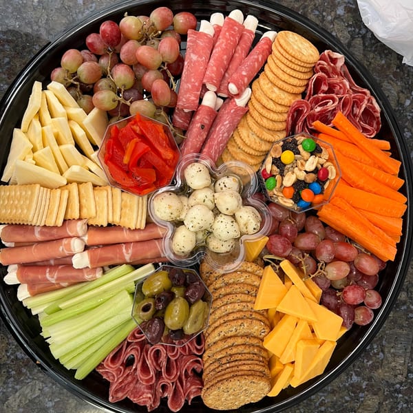 A large black platter filled with various cheeses, meats, crackers, grapes, vegetables, and snacks.