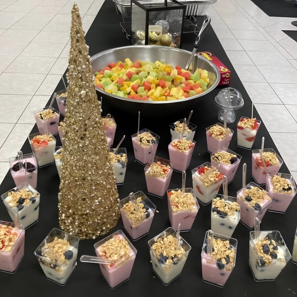 A buffet table with a large bowl of fruit salad, individual yogurt parfaits, and a golden Christmas tree.