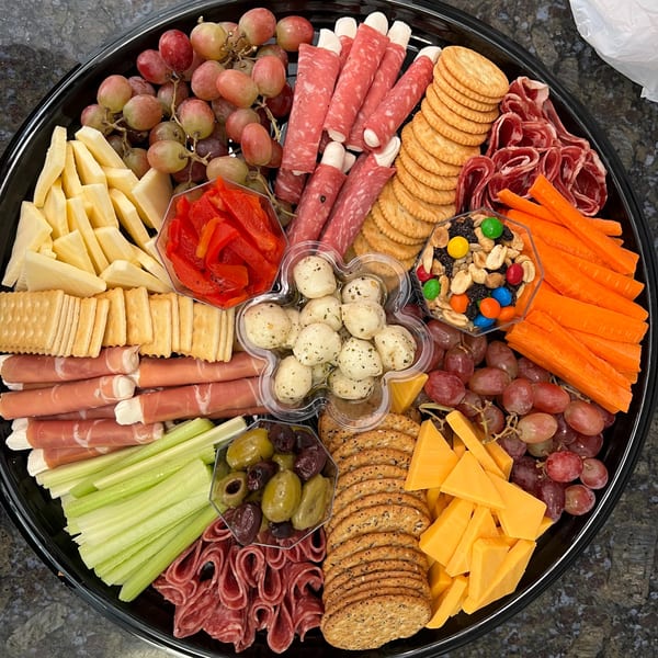 A large black platter filled with various cheeses, meats, crackers, grapes, vegetables, and snacks.